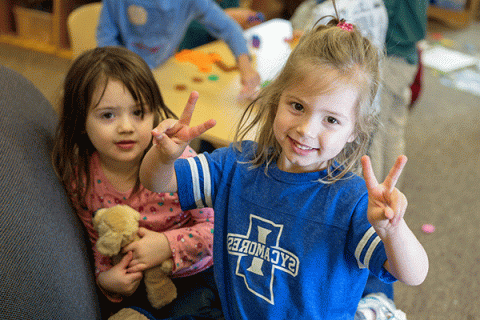 A young blonde girl wearing an Indiana State University shirt smiles at the camera in the Early Childhood Learning Center as a young dark-haired girl beside her looks on. Other children are partially visible around a table in the blurred background.